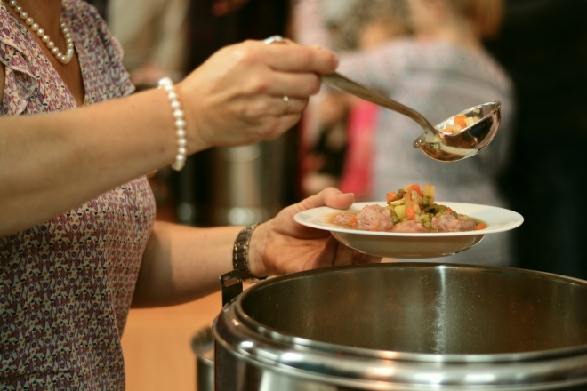 Eine frau hält einen Suppenteller in der Hand und schaufelt mit der anderen Hand Suppe aus dem Topf auf den Teller. 