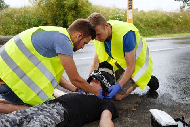 Motorradunfall. Person mit einem Helm liegt am Boden. Neben der Person knien zwei Ersthelfer und nehmen dem Unfallopfer den Helm ab.