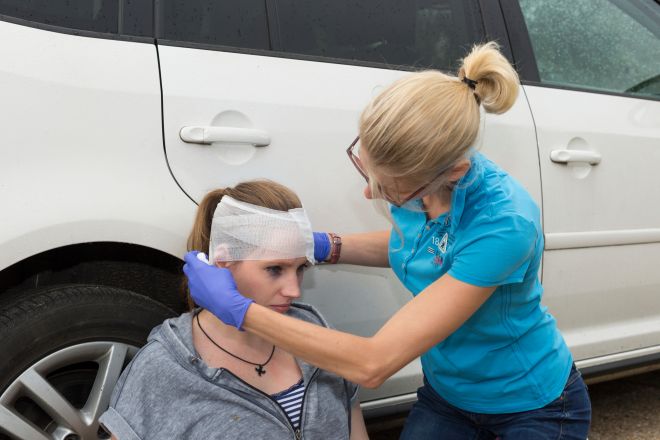 Frau sitzt an einem Auto gelehnt auf dem Boden. Vor ihr kniet eine andere Frau, eine Ersthelferin. Sie legt einen Kopfverband bei der sitzenden sitzenden Person an.