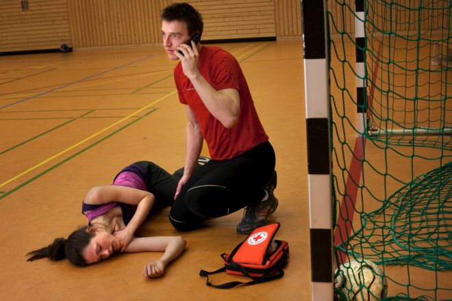 Unfall beim Sport: Junge Frau liegt auf dem Fußboden in einer Sporthalle. Sie ist in die stabile Seitenlage gebracht worden. Der Mann neben ihr fordert telefonisch weitere Hilfe an. Neben ihnen liegt ein Verbandset mit dem DRK-Logo.