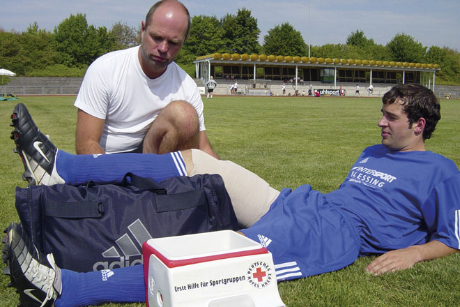 Ein Spieler hat sich beim Fußballspielen verletzt. Er liegt auf dem Feld und hat sein Bein hoch gelagert. Als Erhöhung dient hierfür eine Sporttasche. Der Oberschenkel des hochgelagerten Beines ist bandagiert. Neben dem aktiven Sportler kniet ein Mann. Zudem ist noch eine Kühltasche mit dem DRK-Logo zu sehen. 