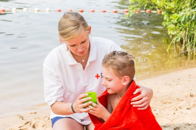 Szene am See: Frau kniet am Strand bei einem Kind, was in eine Decke gehüllt ist. 