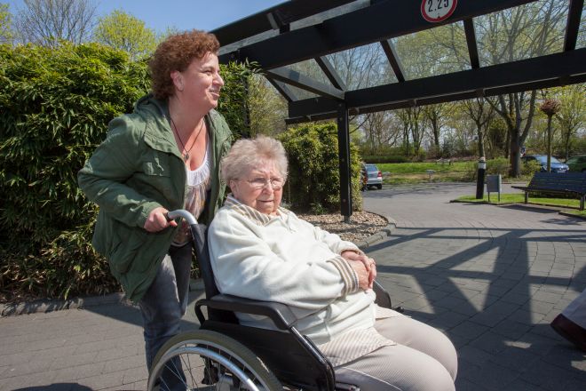 Eine Frau schiebt eine Seniorin im Rollstuhl an der frischen Luft.