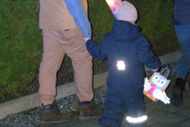 Kinder in einem dunklen Anzug trägt eine selbstgebastelte Laterne in der Hand. Da Motiv entspricht einer Eule. Das Kind läuft an der Hand eines Angehörigen.