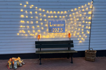 Eine Holzbank steht vor einer Wand. Dahinter ist eine Lichterkette an der Wand befestigt mit einem Schild. Aufschrift Lichterfest 2023. 