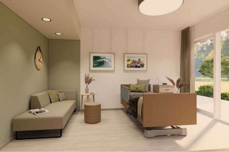 Blick in ein Pflegezimmer. Der gerade Blick richtet sich auf das Bett an der Wand. Links ist eine farbige Wand mit einem Sofa davor platziert. Rechte Hand ist die Fensterfront zu sehen. 