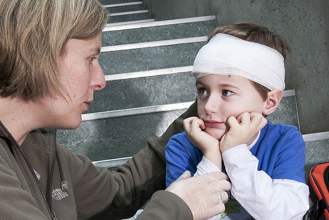 Kleiner Junge sitzt mit einem Kopfverband auf der Treppe. Neben ihm sitzt eine Frau und scheint ihn zu trösten.