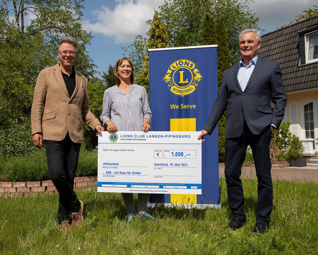 Gruppenbild von drei Personen. Eine Frau steht in der Mitte - zwei Männer links und rechts daneben. Alle Personen halten einen Spendenscheck hoch. Im Hintergrund ein Banner mit dem Logo des Lions Clubs. 