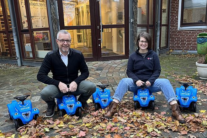 Vor dem Haupteingang der Kita Storchennest sind fünf Bobbycars aufgebaut. Auf zwei der Fahrzeugen sitzen Personen, Detlef Hajann und Nina Neumann.