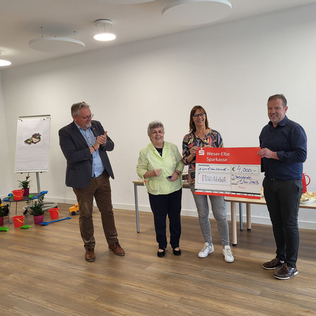 Vier Personen (Mann, Frau, Frau, Mann) stehen in einem Raum nebeneinander und die zwei rechten Personen halten einen Scheck in den Händen. Alle Personen lächeln. 