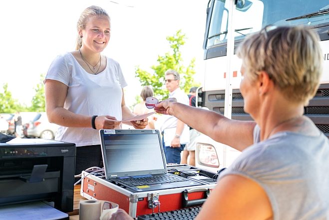 Vor einem Blutspendemobil, bei herrlichem Wetter, steht eine junge Frau mit blonden Haaren und will sich zur Blutspende anmelden.